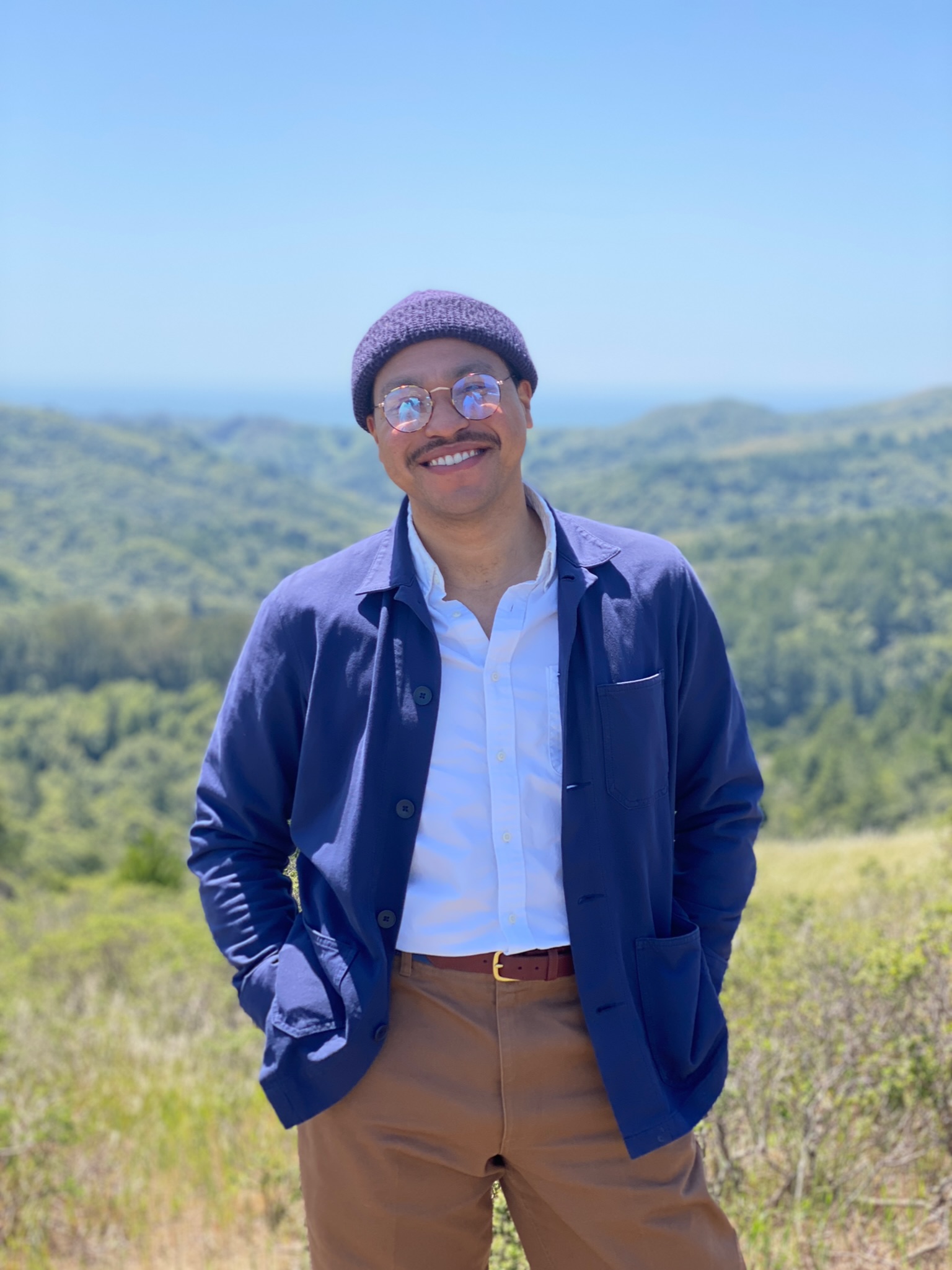 Elijah stands smiling in a beanie, glasses, white button down, khaki pants and a jacket in front of a green mountain background
