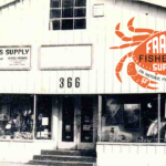 Black and white photo of a storefront with signage reading "Frank's Fisherman's Supply" and a large crab logo on the facade. The building has two large front windows displaying various fishing supplies.